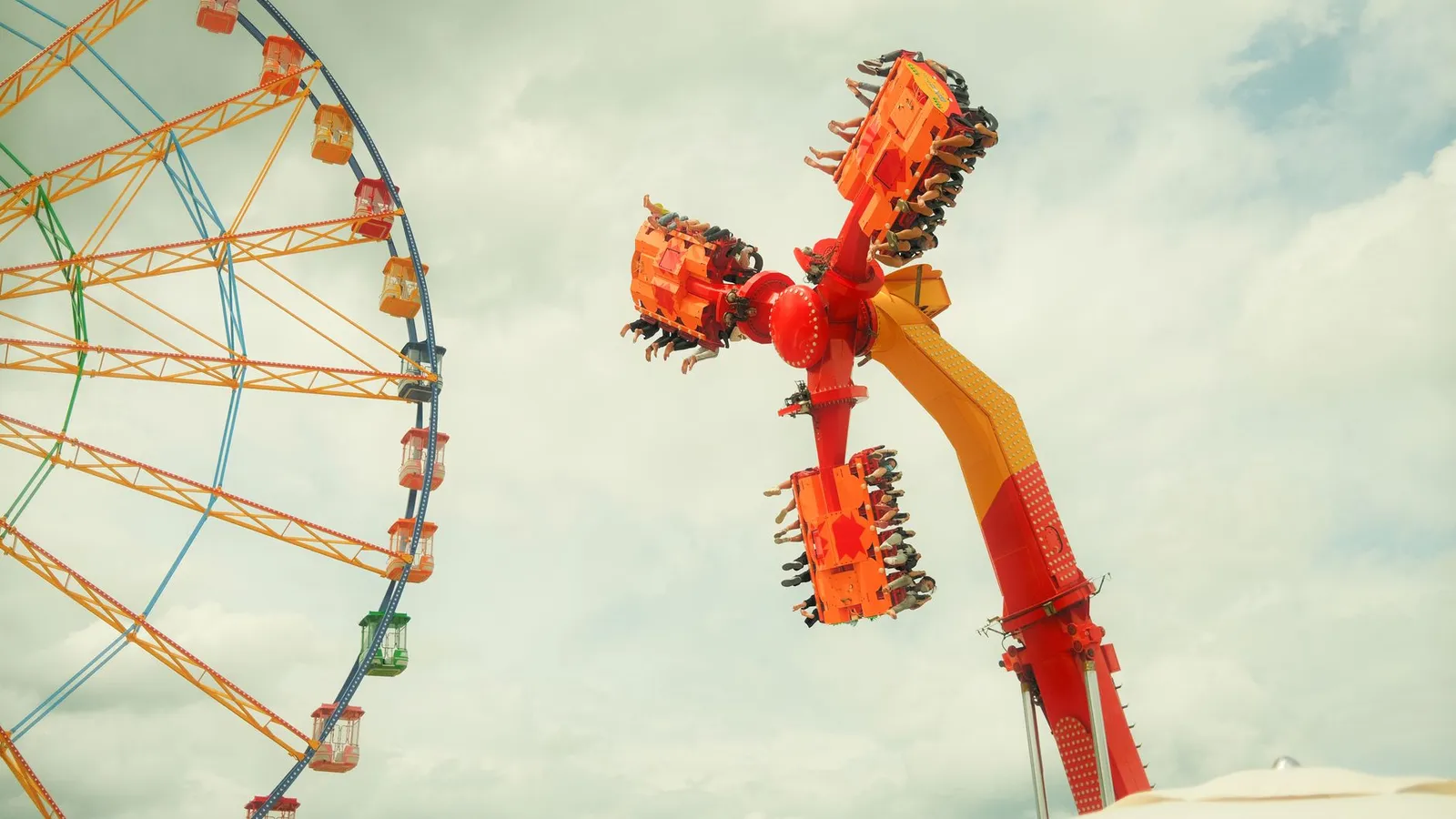Exciting view of a thrilling amusement park ride next to a colorful Ferris wheel under cloudy skies.