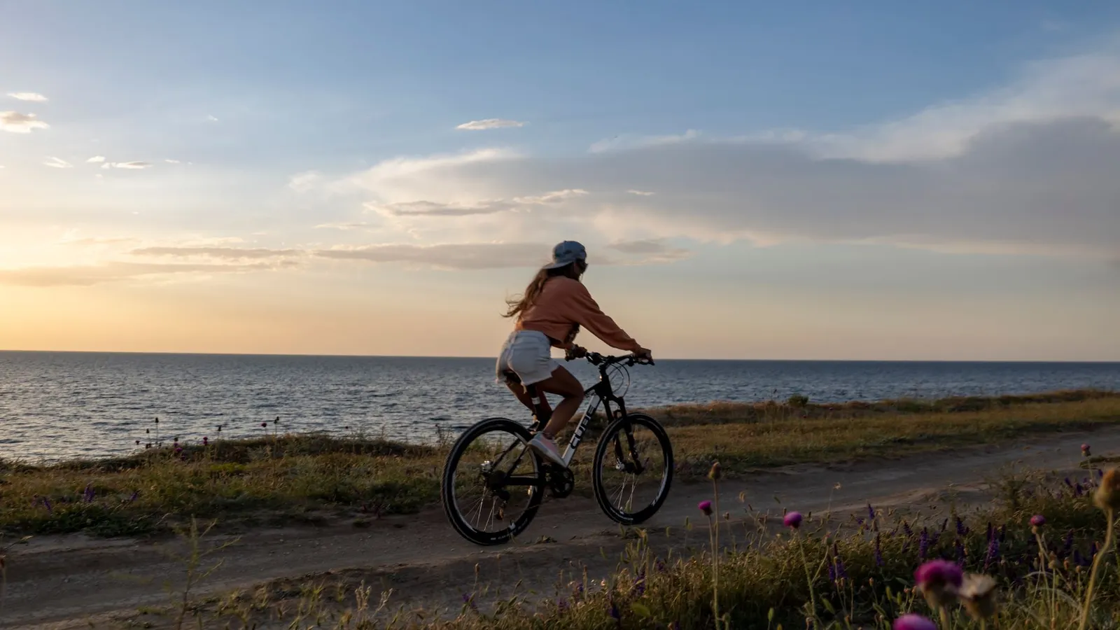 A woman rides her bicycle along a seaside path under a serene sunset, embracing nature.