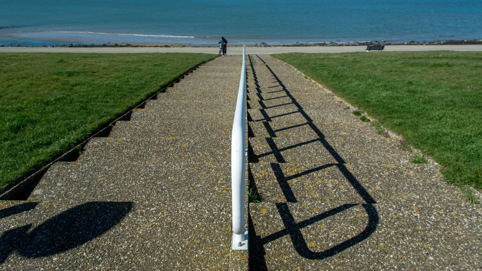 A peaceful coastal scene featuring a pathway, ocean view, and distant cyclist on a sunny day.