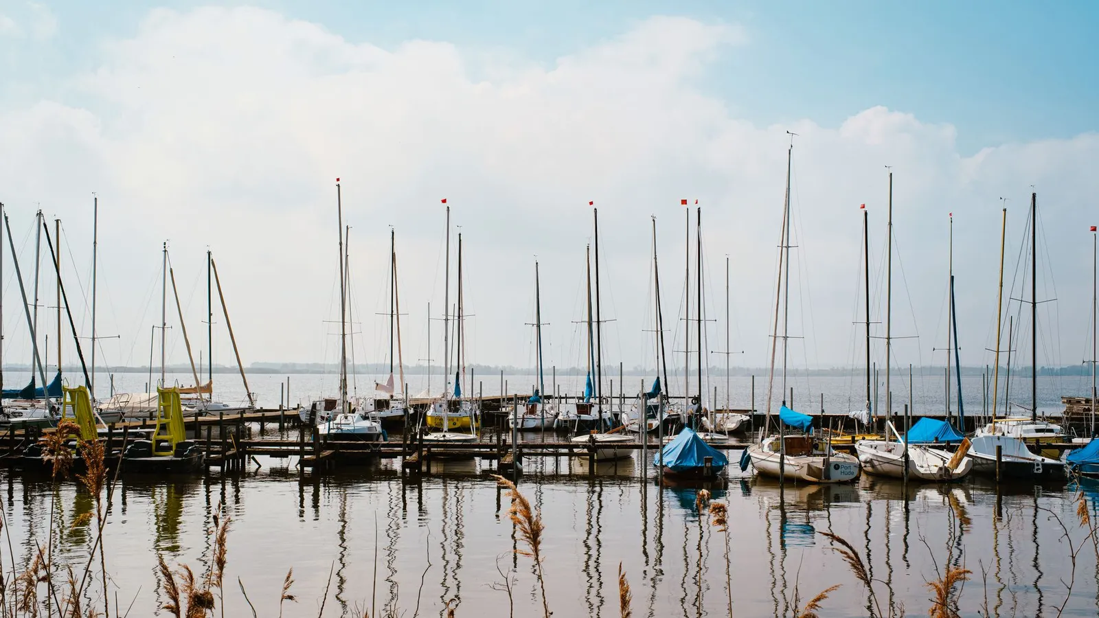 Peaceful harbor scene with sailboats moored under a bright blue sky, reflected on calm waters.