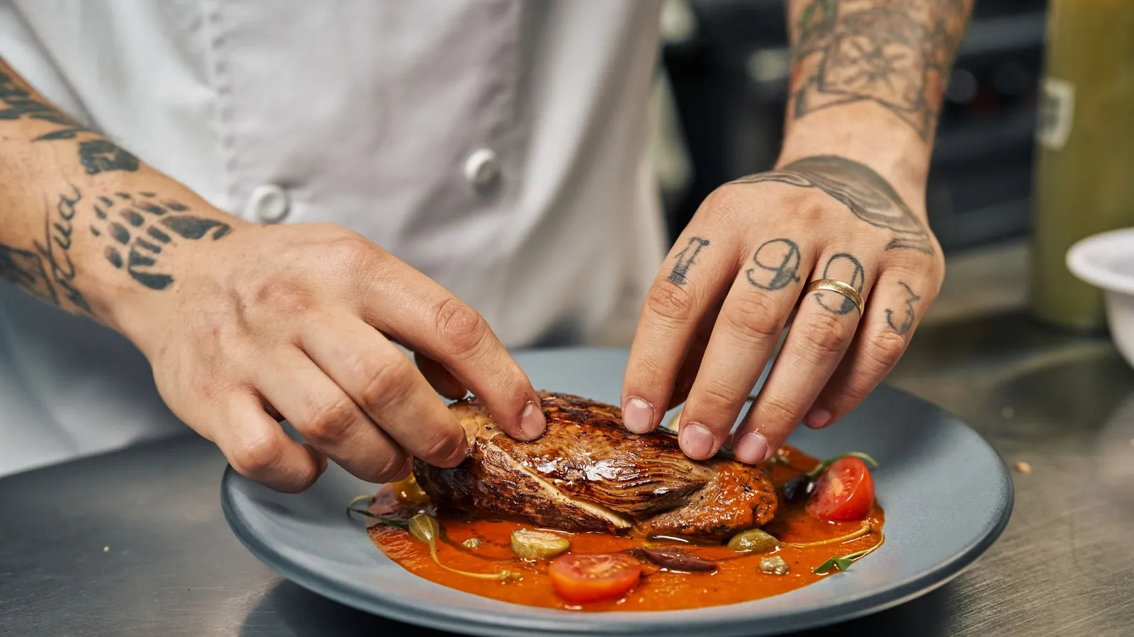 Chef with tattoos presenting a perfectly cooked steak with garnishes on a colorful plate.
