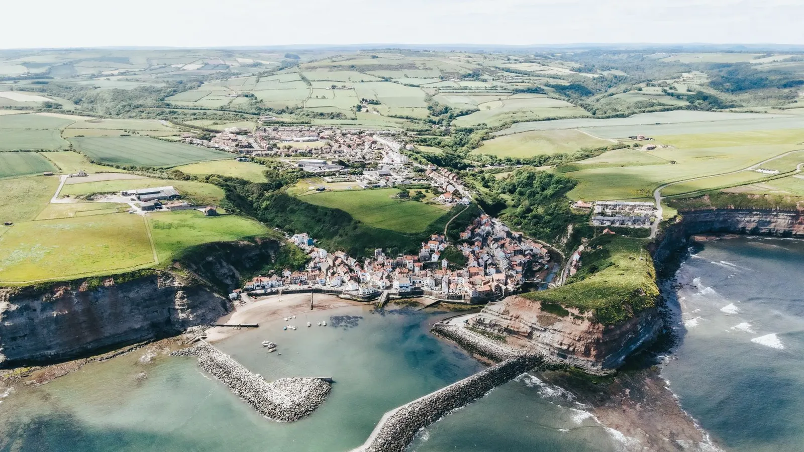 Dramatic aerial shot of a coastal village surrounded by cliffs and green fields.