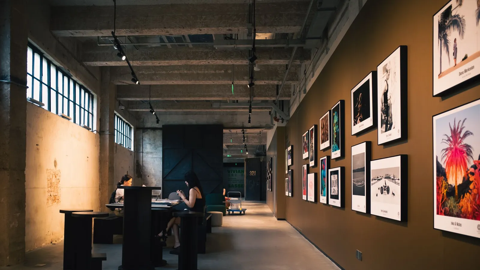 Art gallery hallway with framed artworks and visitors seated inside, under warm lighting.