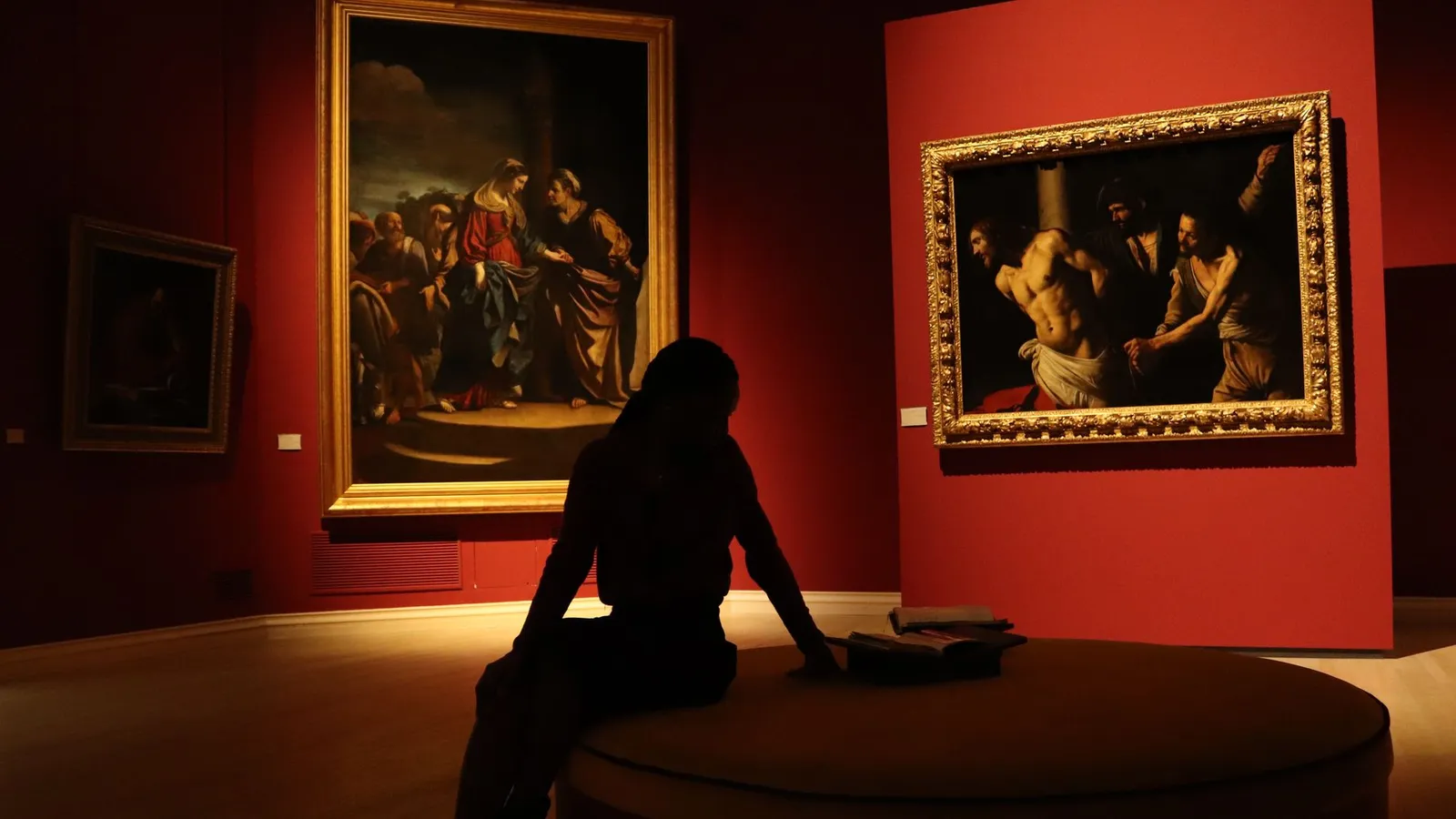 Silhouetted visitor in an art gallery with dramatic lighting on classic paintings.
