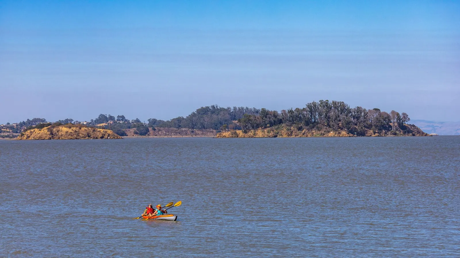 Two kayakers paddling near a scenic island under a clear blue sky.