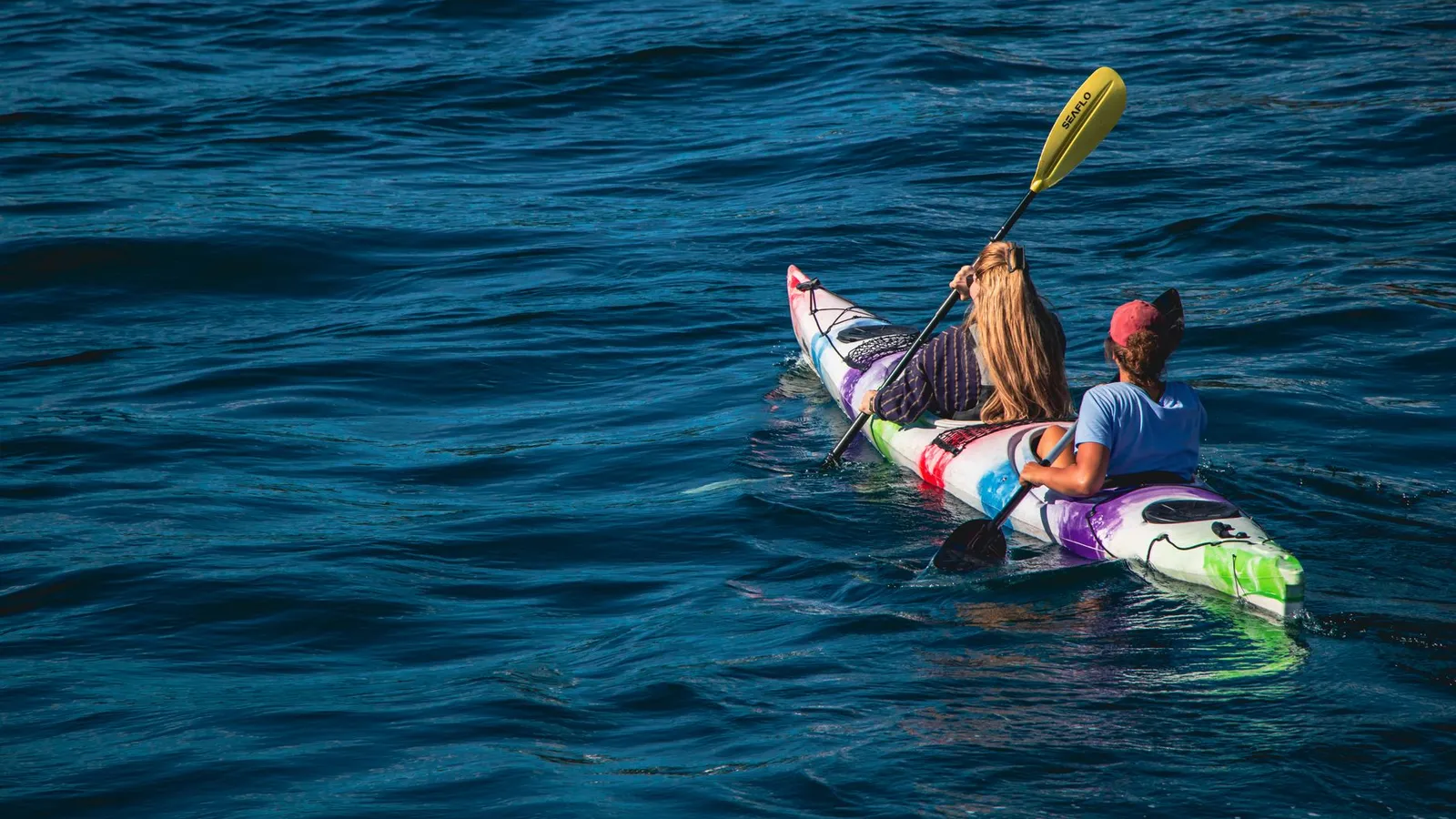 Two women paddle a colorful kayak on a calm lake during a sunny summer day, enjoying outdoor recreation.