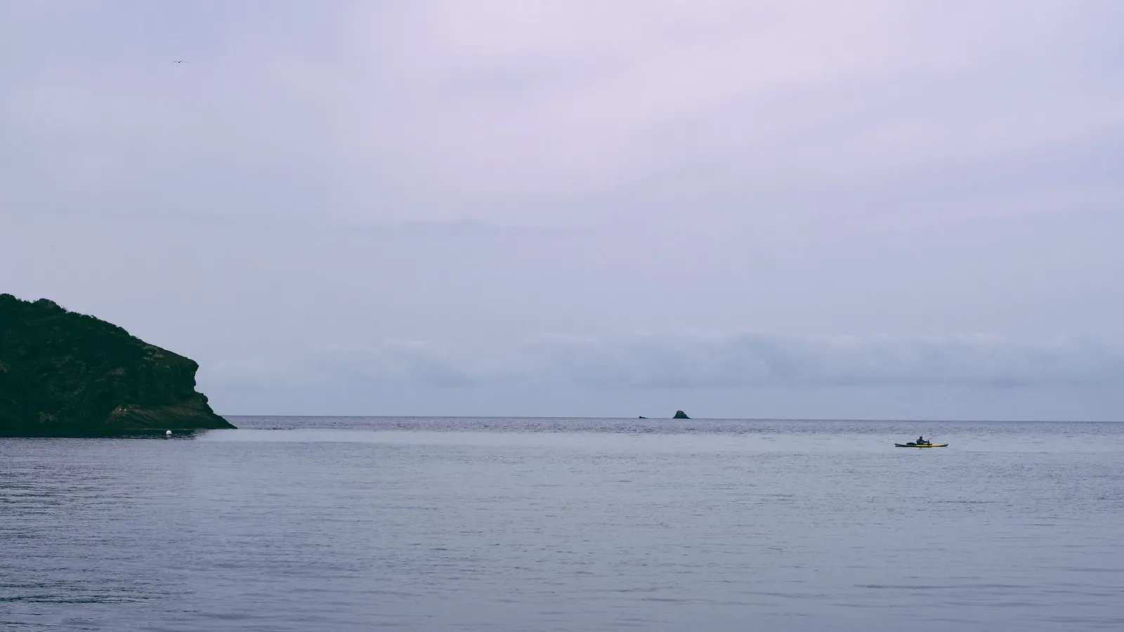 A tranquil coastal scene with a lone kayaker on calm waters near a rugged cliff.