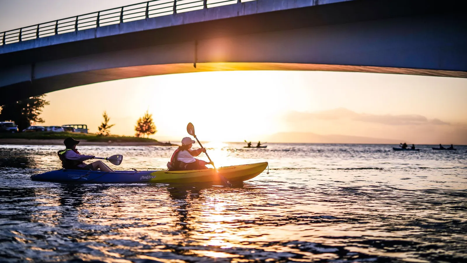 Two kayakers paddle under a bridge as the sun sets, casting warm reflections on the water.
