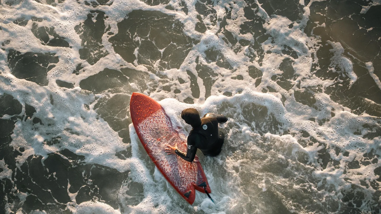 Surfer with red board navigating waves, captured from above.